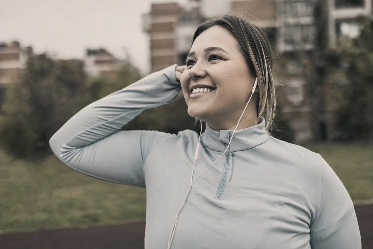 A woman in athletic wear smiling outdoors while putting in an earphone before a run