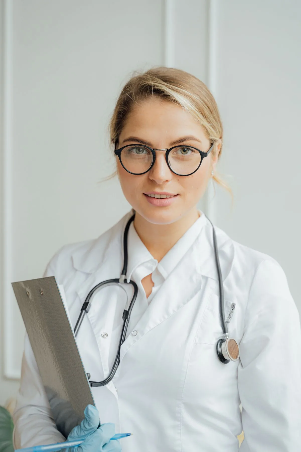 A physician wearing glasses and a white coat with a stethoscope, holding a clipboard and pen with gloved hands