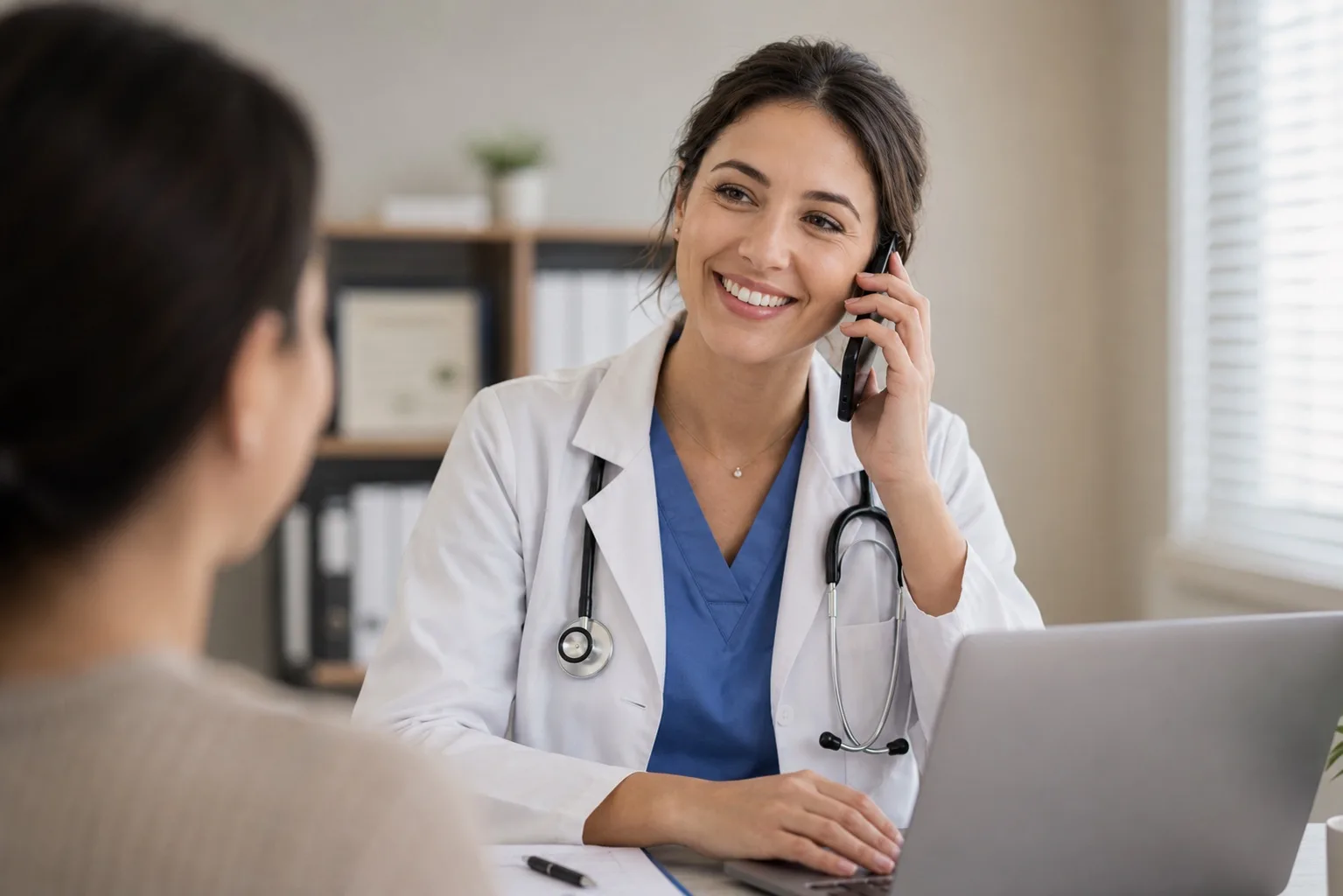 A smiling physician in a white coat taking a phone call at her desk, laptop open in front of her