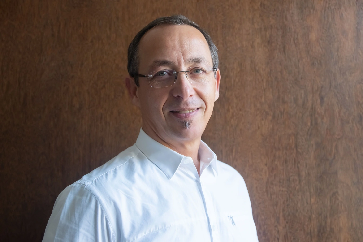 A man in his fifties with rimless glasses and a white open-collar shirt, smiling slightly toward the camera in front of a warm wood-panelled wall