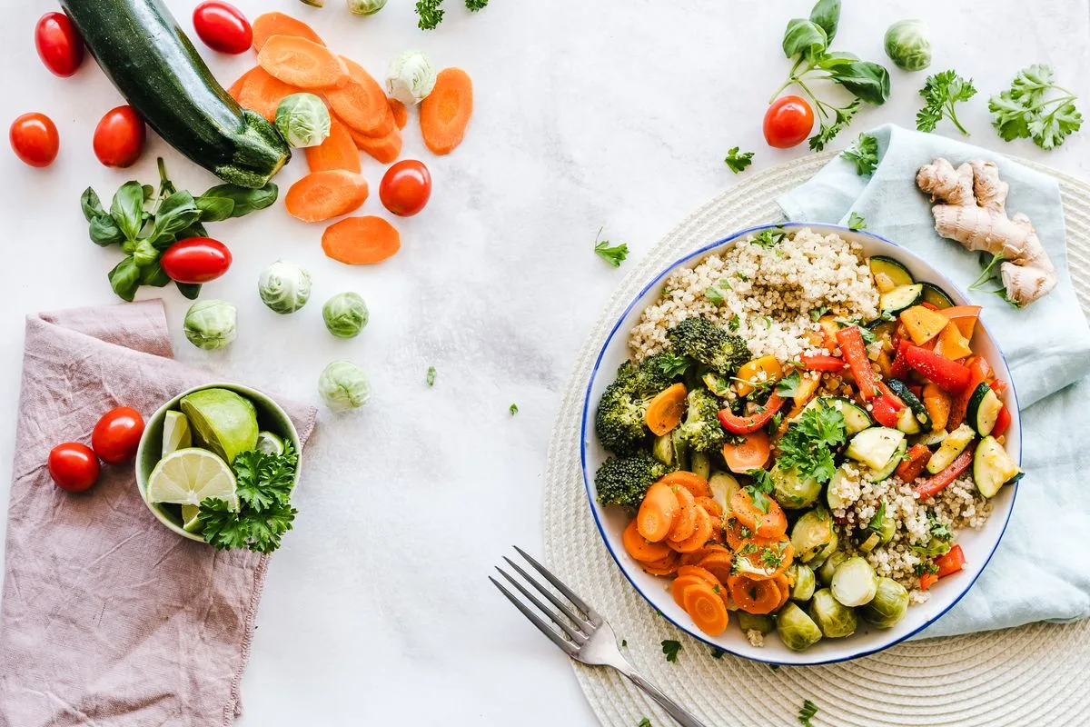 Overhead view of a quinoa and roasted vegetable bowl with broccoli and carrots, surrounded by cherry tomatoes, brussels sprouts, basil, ginger and lime wedges on a white countertop