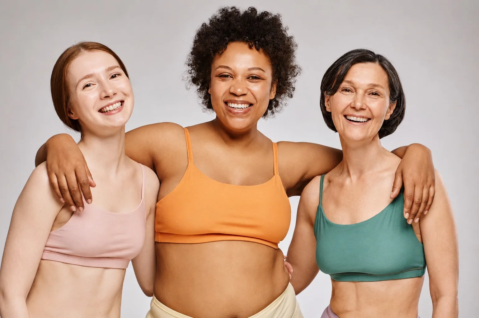 Three women of different ages and body types in athletic wear, smiling with their arms around each other