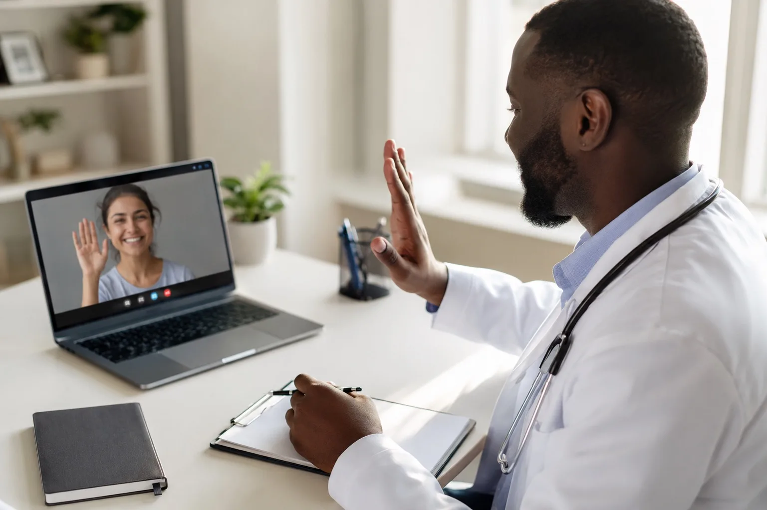 A physician in a white coat waving to his patient at the start of a video visit on his laptop